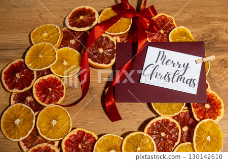 MERRY CHRISTMAS text on postcard with red envelope on dried slices of grapefruit and oranges in Christmas wreath. Zero waste eco-friendly Christmas. Sustainable greeting card MERRY CHRISTMAS text on postcard with red envelope on dried slices of grapefruit and oranges in Christmas wreath. Zero waste eco-friendly Christmas. Sustainable greeting card 130142610