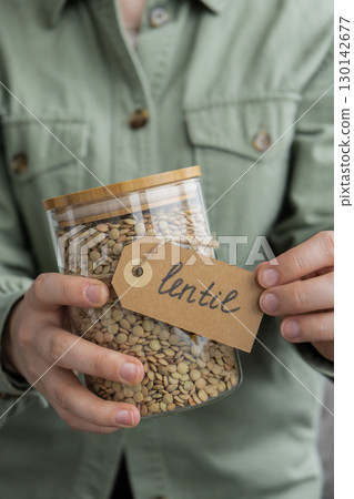 Unrecognizable woman holding refillable glass jar with raw green lentil porridge. Refill product format eco-friendly living. Filling reusable container Whole spelt grain species. High protein fiber 130142677