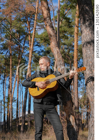 Beautiful blonde man playing acoustic guitar and singing relaxing enjoying outdoors on sunny autumn day. Artist learning and practicing playing musical instrument. Fall leaves autumnal 130142690