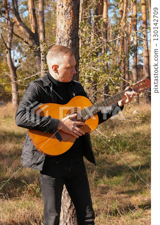 Beautiful blonde man playing acoustic guitar and singing relaxing enjoying outdoors on sunny autumn day. Artist learning and practicing playing musical instrument. Fall leaves autumnal Beautiful blonde man playing acoustic guitar and singing relaxing enjoying outdoors on sunny autumn day. Artist learning and practicing playing musical instrument. Fall leaves autumnal 130142709