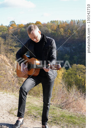 Blonde man playing string guitar outdoors in autumn forest. Concept of sound therapy, mental health and wellness rituals. Calmness tranquility audio-sensory practices. Aura farming energy 130142710