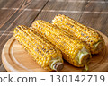 Three grilled corn cobs with char marks resting on wooden cutting board 130142719