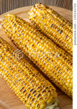 Three grilled corn cobs with char marks resting on wooden cutting board 130142720
