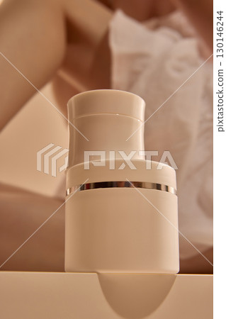 White cosmetic bottle with silver band on table in front of woman body in soft fabric 130146244