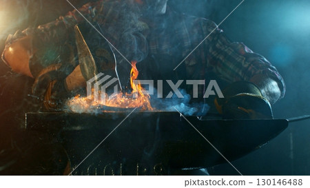 The blacksmith manually forging the molten metal on the anvil in smithy with spark fireworks 130146488