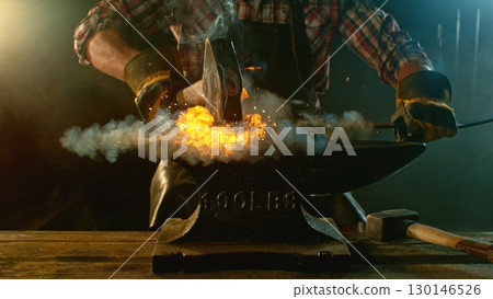 The blacksmith manually forging the molten metal on the anvil in smithy with spark fireworks 130146526