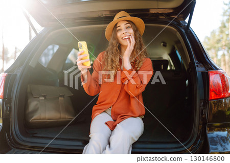 Beautiful woman sits in the trunk of a car outdoor and uses a smartphone. Lifestyle 130146800