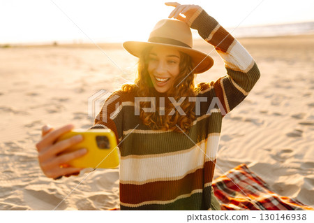Young woman taking a cheerful selfie on a sandy beach during sunset. Lifestyle, travel, active life 130146938