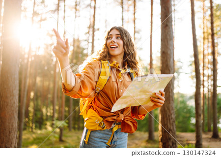 A traveler woman stands in a lush forest, examining a map with a focused expression A traveler woman stands in a lush forest, examining a map with a focused expression 130147324