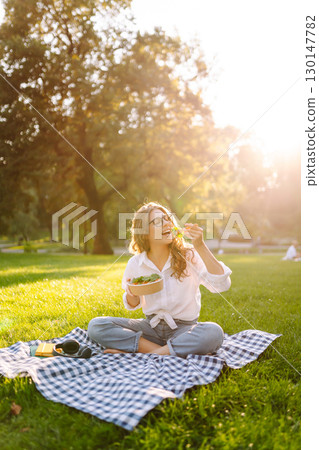Woman enjoying a poke bowl on sunny day in a park while sitting on a picnic blanket. Concept picnic Woman enjoying a poke bowl on sunny day in a park while sitting on a picnic blanket. Concept picnic 130147782