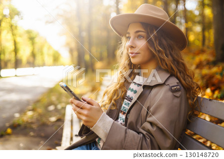 Young smiling woman sitting on bench in autumn park using phone in stylish clothes happy mood. 130148720