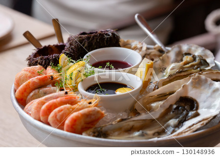 Seafood (oysters, shrimps, sea-urchins, mussels) are decoratively put on ice in a big plate Seafood (oysters, shrimps, sea-urchins, mussels) are decoratively put on ice in a big plate 130148936