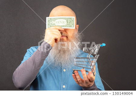 bald man with a large white beard holds a dollar and a model of a grocery basket 130149004
