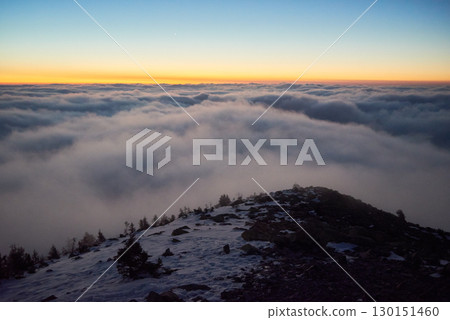 Stunning landscape photograph captures serene beauty of snowy mountain peak at dawn, with breathtaking sea of clouds stretching into horizon. Sky transitions from deep blue to warm orange hue. 130151460