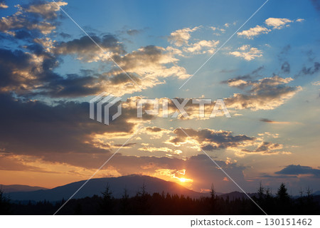 Serene evening sky with hues of blue and soft pink clouds as sun sets. Horizon glows gently, casting light over silhouetted pine trees and rolling hills, creating peaceful and expansive landscape. 130151462