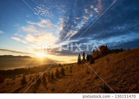 Sun rises over mountains, casting golden rays across landscape. Sky filled with dramatic clouds, creating stunning contrast with clear blue above. Warm light illuminates rolling hills and pine trees. 130151470
