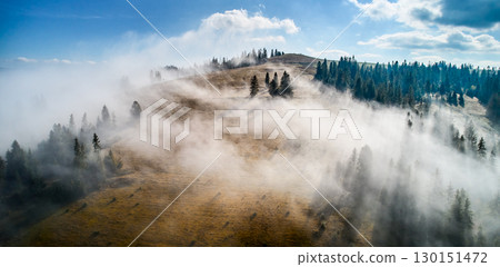 Stunning landscape with dense forests and rolling hills partially shrouded in soft, ethereal fog. Vibrant autumn colors of trees contrast beautifully with clear blue sky and distant mountain peaks. 130151472