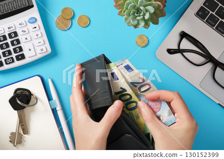 Hands taking euro banknotes out of a wallet. Around them are a calculator, coins, a laptop, and glasses. Managing cash and personal finances. 130152399