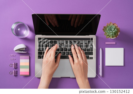 Hands typing on laptop keyboard with stationery, plant, and glass of water on purple desk. Idea of freelancing, learning, working from home. Hands typing on laptop keyboard with stationery, plant, and glass of water on purple desk. Idea of freelancing, learning, working from home. 130152412