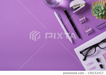 Workspace with a laptop, pen, stapler, glasses, paper clip, plant, and glass of water on a purple background. Space for your text. 130152417