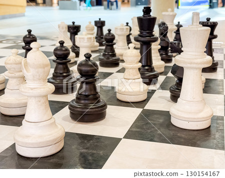 Large chess pieces on outdoor board arranged for a game. Strategy, logic, and intellectual challenge in symbolic black and white setting. 130154167