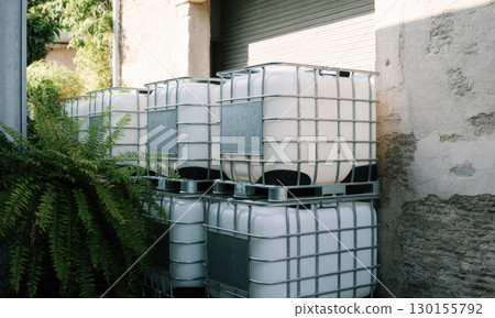 Stacked white IBC containers with metal cages and green foliage in an industrial setting 130155792