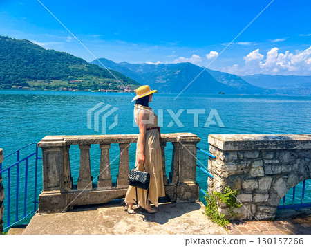 Woman in Maxi Dress and Straw Hat Gazes at a Stunning Mountain-Rimmed Lake Below Woman in Maxi Dress and Straw Hat Gazes at a Stunning Mountain-Rimmed Lake Below 130157266