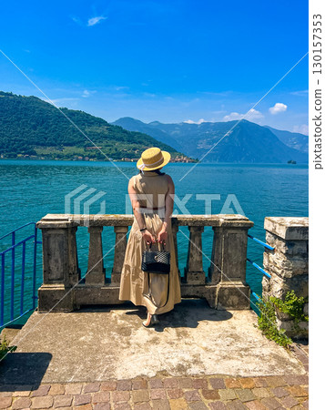 Woman in Maxi Dress and Straw Hat Gazes at a Stunning Mountain-Rimmed Lake Below Woman in Maxi Dress and Straw Hat Gazes at a Stunning Mountain-Rimmed Lake Below 130157353