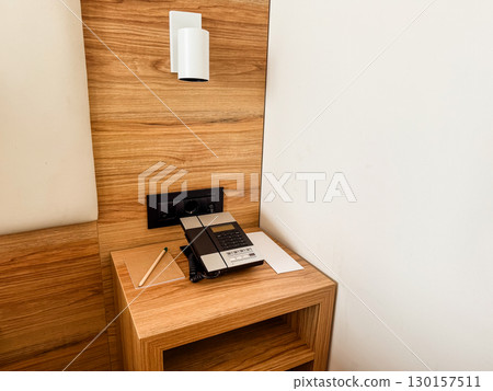 Bedside table with telephone, lamp, and notepad in hotel room. Communication, service, and assistance concept in hospitality environment for guest comfort. Bedside table with telephone, lamp, and notepad in hotel room. Communication, service, and assistance concept in hospitality environment for guest comfort. 130157511
