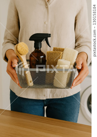 Person holding stylish mesh basket filled with eco friendly cleaning supplies wooden brush glass spray bottle and natural sponges, conveying sense of care and cleanliness 130158184