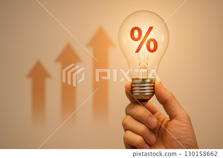Hand holding glowing light bulb with red percentage symbol inside and rising upward arrows in blurred background, symbolizing investment growth and interest rate increase 130158662