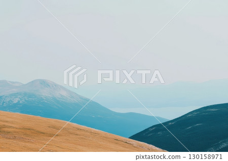 Mountain landscape with rolling hills and distant water view under a hazy sky during daylight Mountain landscape with rolling hills and distant water view under a hazy sky during daylight 130158971