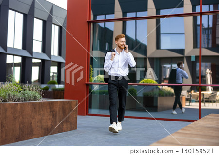 Bearded redhead man walking near glass facade talking on smartphone 130159521