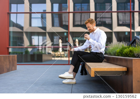 Businessman sitting on bench enjoying coffee to go and scrolling via digital tablet 130159562
