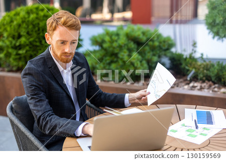 Ginger-haired man in suit reading reports while using laptop outdoor Ginger-haired man in suit reading reports while using laptop outdoor 130159569