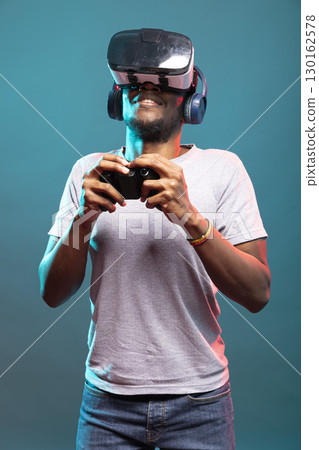Young man playing a video game with VR headset and controller, fully immersed in futuristic simulation. Male model with wireless headphones, holding a gamepad and enjoying a virtual reality interface. Young man playing a video game with VR headset and controller, fully immersed in futuristic simulation. Male model with wireless headphones, holding a gamepad and enjoying a virtual reality interface. 130162578
