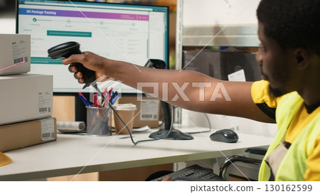 Male wheelchair user in hi vis vest scanning barcode shipping labels, processing cargo orders on pc. Warehouse clerk handles online shopping operations for retail large business. Camera B. 130162599