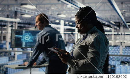 Data center system administrator uses AI on tablet to help customers resolve website hosting problems. African american woman in server farm helps users keep online services running smoothly, camera B 130162602