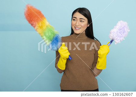 Happy asian woman wearing yellow rubber gloves holds rainbow and white duster, smiling confidently while posing for portrait against blue background. Housekeeping and hygiene concept. 130162607
