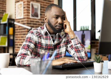 Bearded male manager works from home, looking frustrated and bored while checking project updates on computer. Self employed black man sits in living room overwhelmed by digital tasks. Bearded male manager works from home, looking frustrated and bored while checking project updates on computer. Self employed black man sits in living room overwhelmed by digital tasks. 130162629