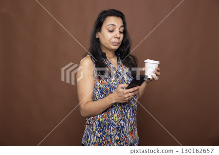 Indian woman stands against brown background, holding her coffee and using smartphone. Female model casually holds takeaway cup and scrolls through her mobile device, engaged in online conversation. 130162657