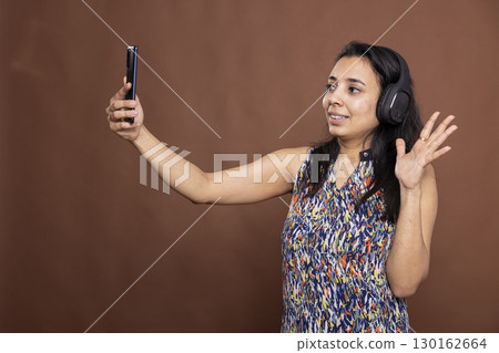 Relaxed indian woman with wireless headphones holding mobile phone, smiling and waving at screen. Cheerful female individual having video call with family, standing against brown background. Relaxed indian woman with wireless headphones holding mobile phone, smiling and waving at screen. Cheerful female individual having video call with family, standing against brown background. 130162664