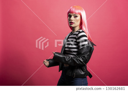 Gorgeous woman with striped crop top stands facing camera under soft lighting with vibrant hairstyle and radiant skin. Young model in black jacket conveys calm elegance, posing against pink background Gorgeous woman with striped crop top stands facing camera under soft lighting with vibrant hairstyle and radiant skin. Young model in black jacket conveys calm elegance, posing against pink background 130162681
