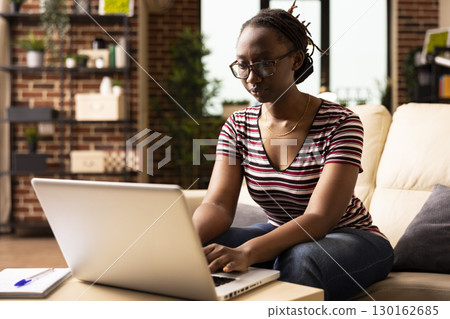 Black woman sitting on couch with concentrated expression, typing on laptop, updating resume and online work profile. Focused female freelancer preparing business presentation on personal computer. 130162685