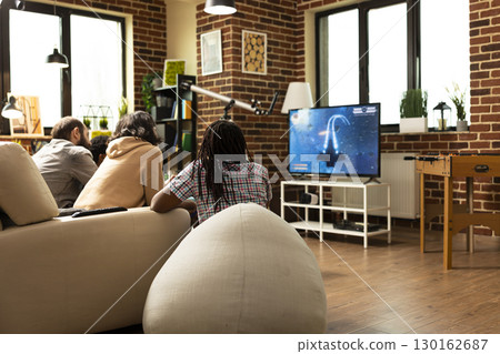 Group of friends enjoy relaxed afternoon at home, facing television screen. Male and female individuals resting on soft furniture and playing videogames, having friendly competition indoors. 130162687