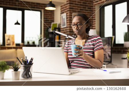 Black female entrepreneur holding mug and working on her personal computer brick wall room. Self employed woman having coffee and using laptop at desk, checking emails and reviewing online documents. 130162688