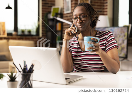 Remote female manager holds cup as she speaks with employee on cellphone, discussing recent company progress. African American woman drinks coffee and answers business calls while working from home. Remote female manager holds cup as she speaks with employee on cellphone, discussing recent company progress. African American woman drinks coffee and answers business calls while working from home. 130162724