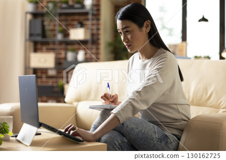 Determined asian woman with notebook and pen, seated in brick wall living room, prepares for investor meeting. Remote female manager reviews company report on laptop and takes strategic notes. Determined asian woman with notebook and pen, seated in brick wall living room, prepares for investor meeting. Remote female manager reviews company report on laptop and takes strategic notes. 130162725
