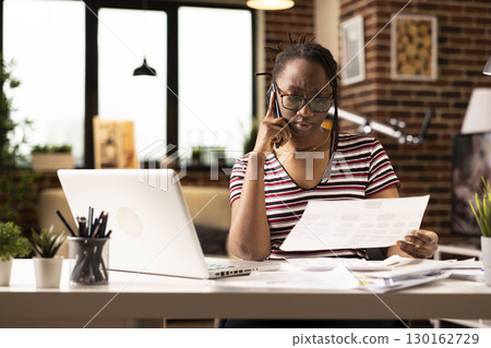 African american freelancer reviewing important papers during serious phone call with business client. Focused black woman with documents in hand, talking with coworker through mobile device. 130162729