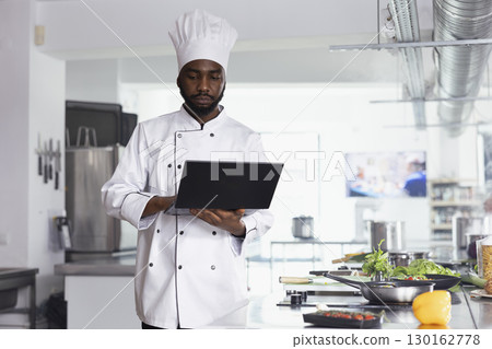 African american chef researching new cooking tips in restaurant kitchen uses a laptop while preparing fresh ingredients and plating food. High quality culinary arts and delicious dishes. African american chef researching new cooking tips in restaurant kitchen uses a laptop while preparing fresh ingredients and plating food. High quality culinary arts and delicious dishes. 130162778